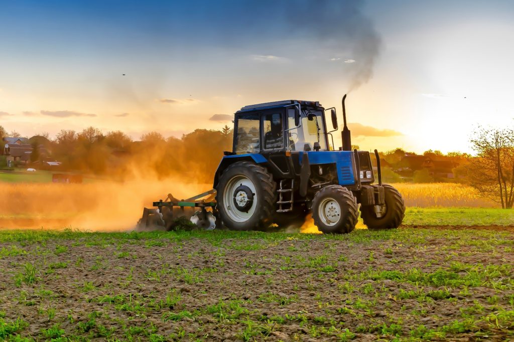 Modern blue tractor machinery plowing agricultural field meadow at farm at spring during sunset